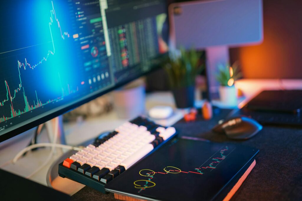 A developer's desk with lines of code on a monitor and cryptocurrency price charts visible.