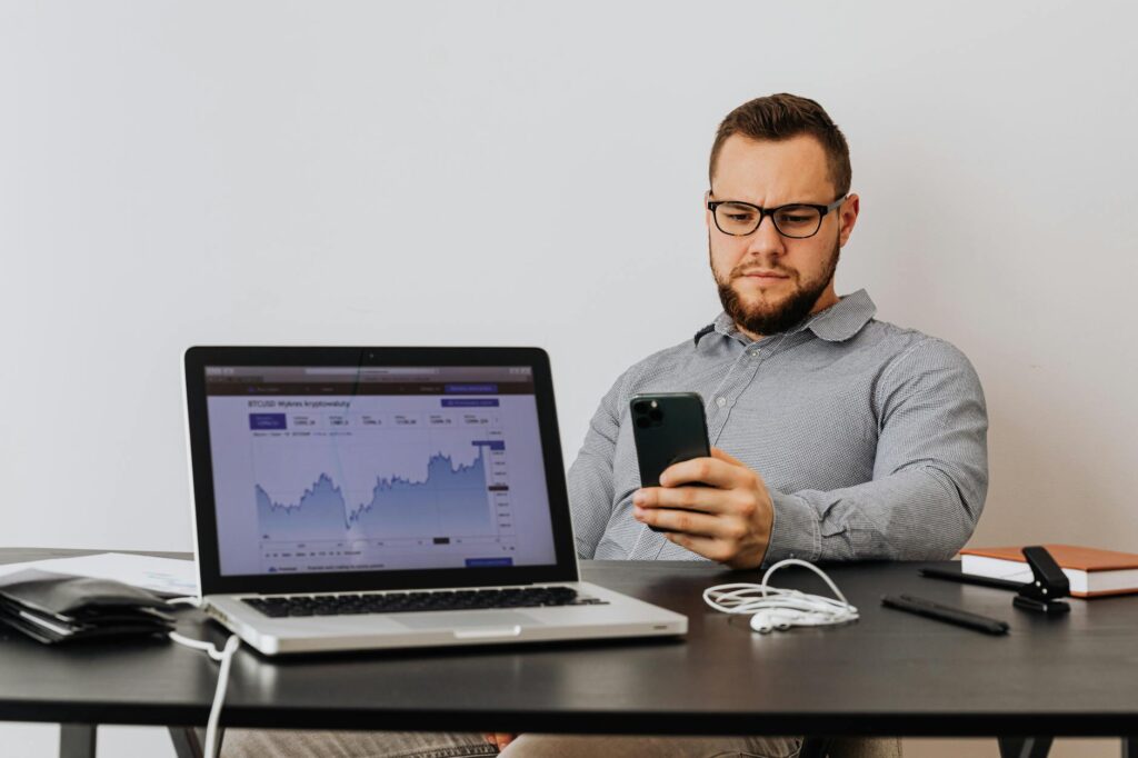 A person holding a smartphone displaying a fluctuating cryptocurrency price chart with green and red candles.