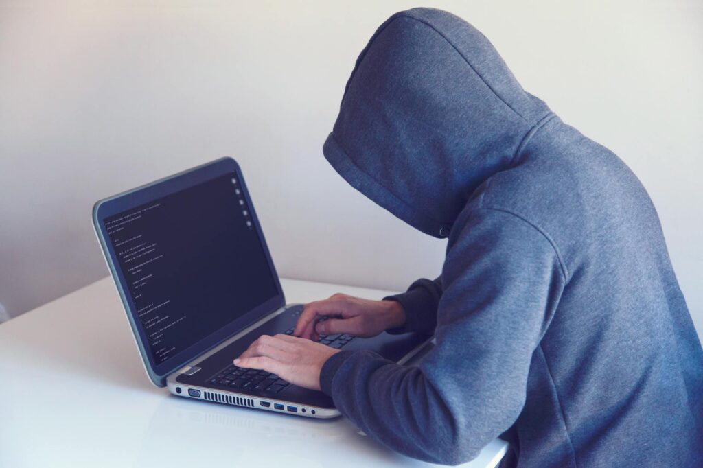 A hooded figure representing a hacker sits in front of a computer with lines of code, illustrating cyber threats.