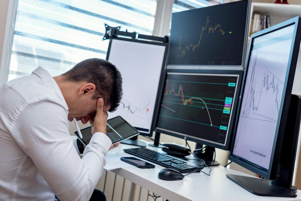 A frustrated investor holding their head in their hands while looking at a volatile crypto price chart on a monitor.