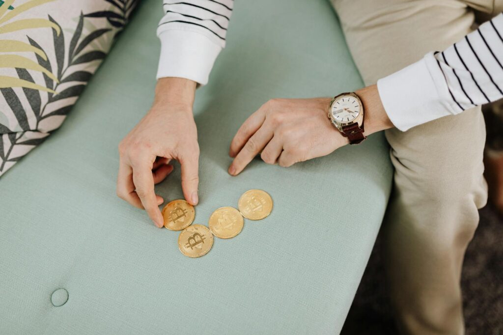 A close-up of a person's hand holding a golden Bitcoin, with a blurred laptop screen showing charts in the background.