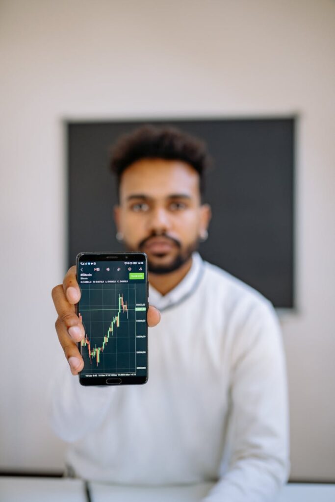 A person with a concerned expression holding a smartphone that shows a declining cryptocurrency price chart.