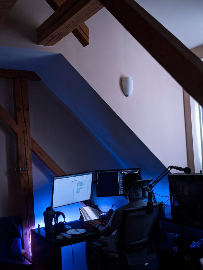 A software developer working in a dark room with multiple monitors displaying lines of code and cryptocurrency price charts.