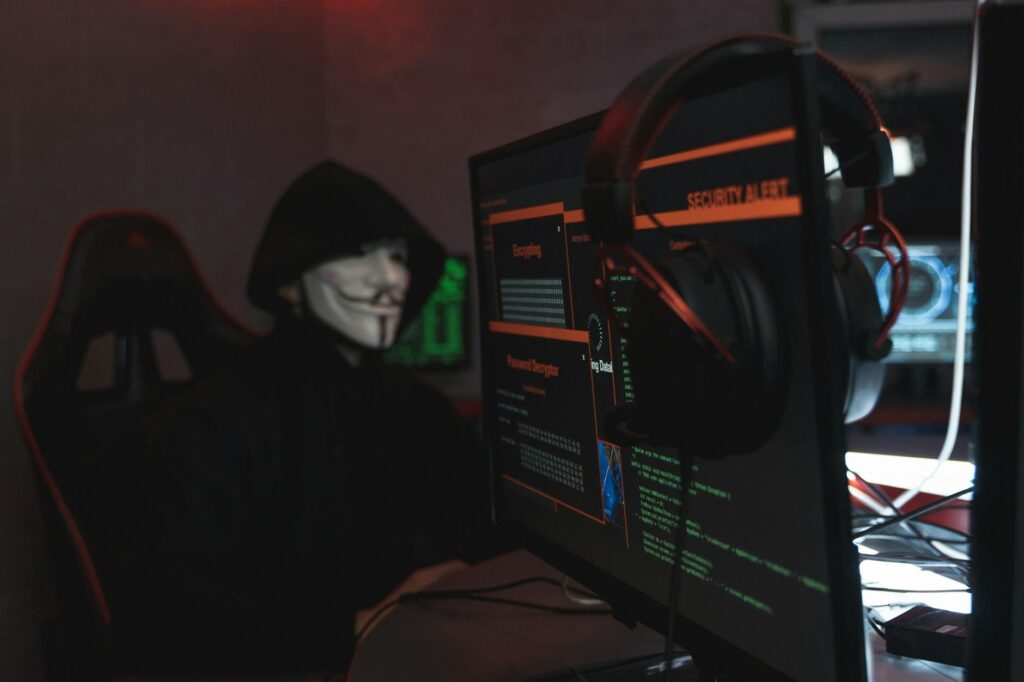 A silhouette of a person in a hoodie sitting in front of multiple computer monitors displaying green code, illustrating a security threat.