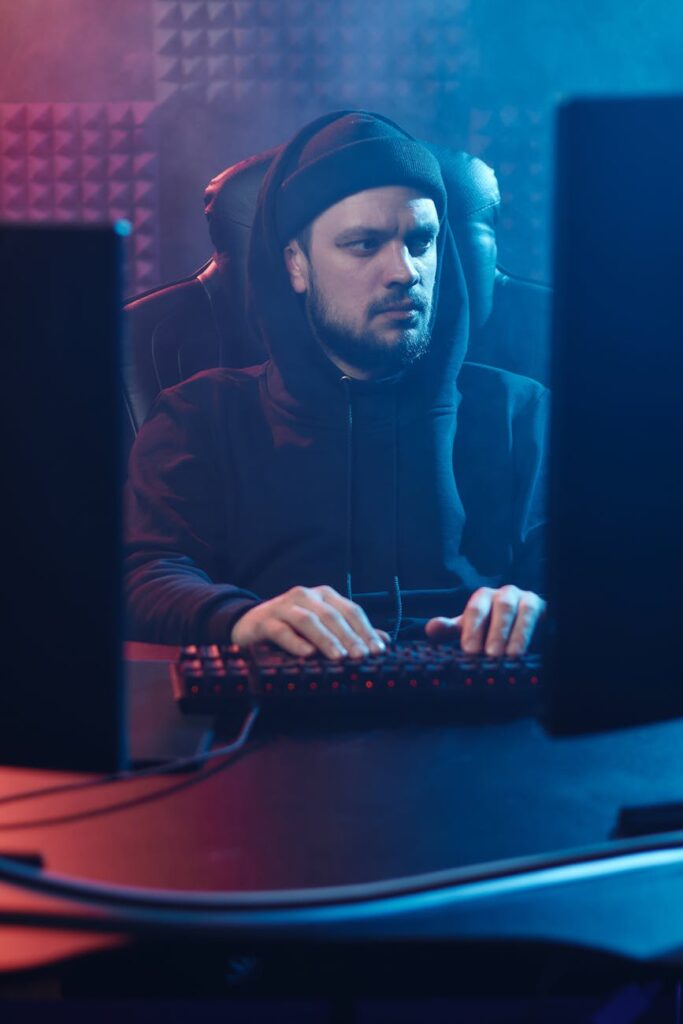 A person in a hoodie sitting in a dark room, with complex code reflected on their face, representing a hacker.